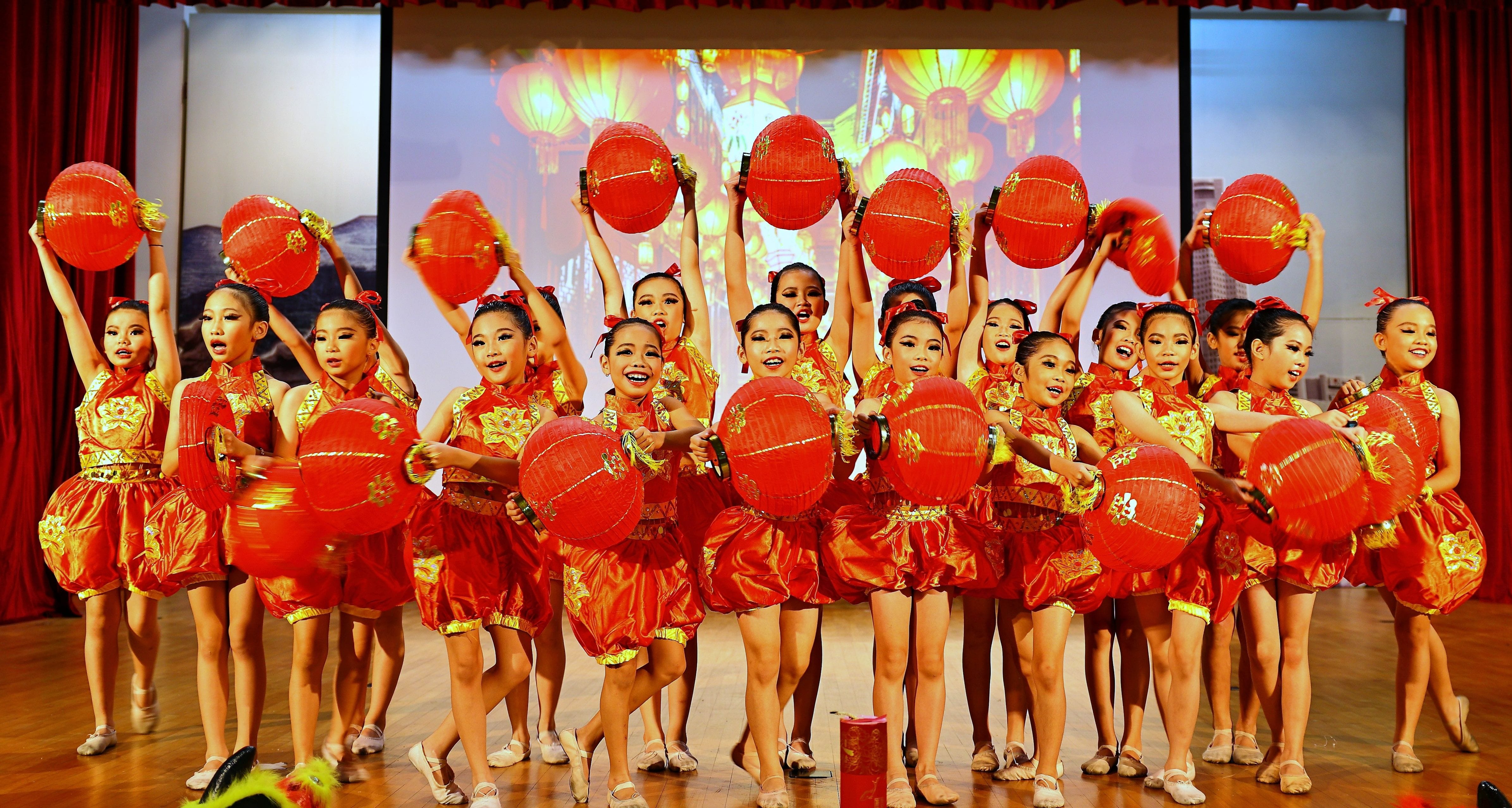 Girls in red and gold costumes hold large red lanterns on stage during a Chinese cultural dance performance.